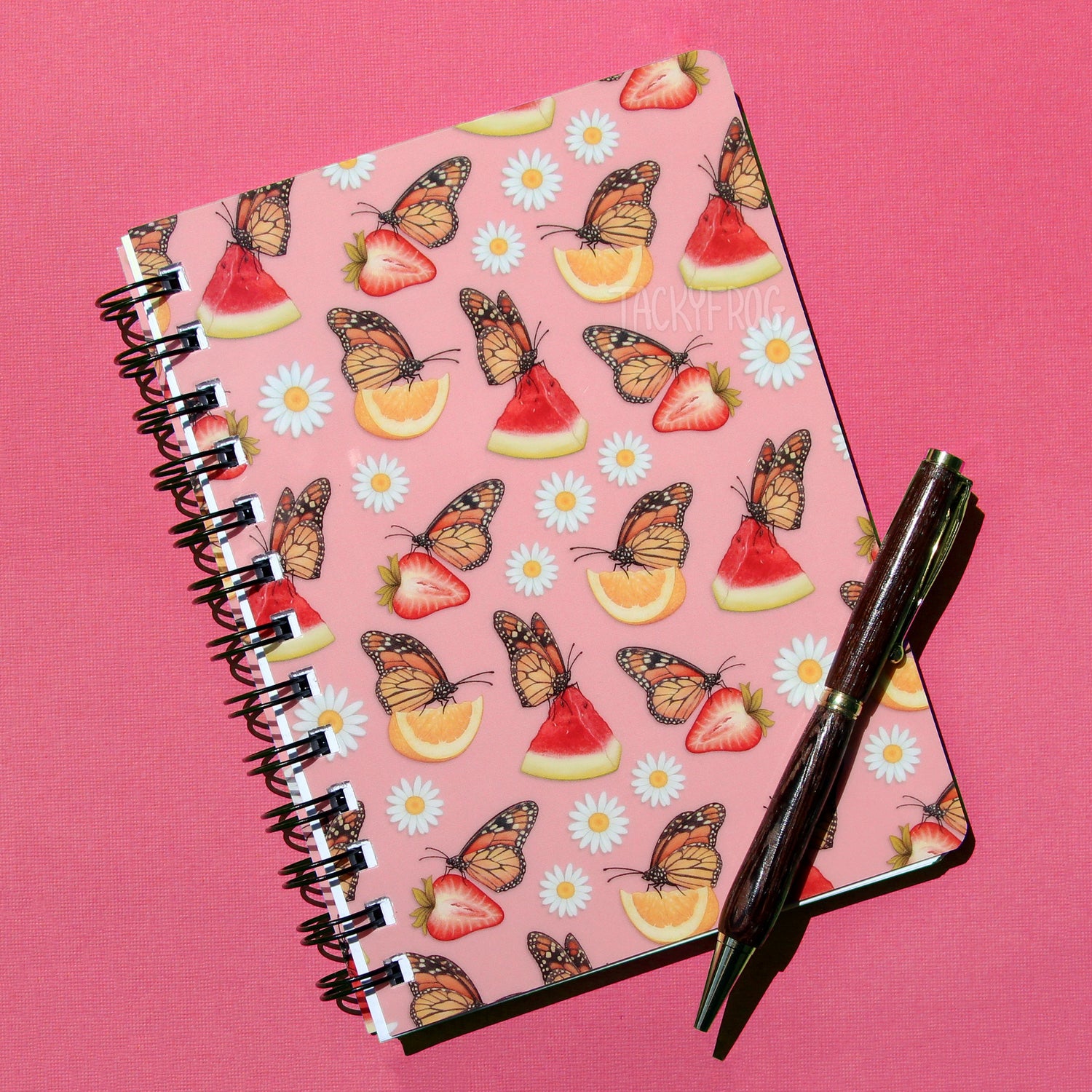 A small pink spiral journal featuring a pattern of white daisies and monarch butterflies on watermelon, strawberry, and lemon slices. The pen is not included.