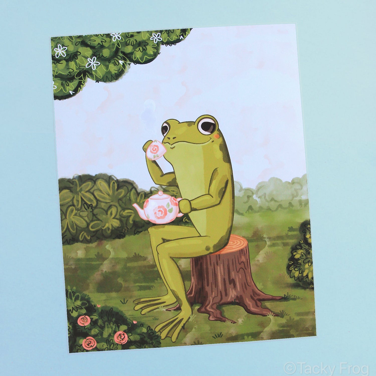 An art print of a green frog drinking tea while sitting on a tree stump in the garden.