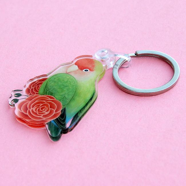 An acrylic keychain depicting a peach-faced lovebird sitting on red flowers