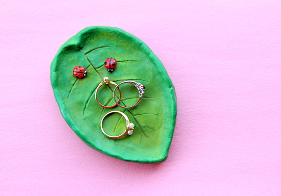 A clay jewelry dish that looks like a leaf with two ladybugs on it. There are three rings inside the dish.