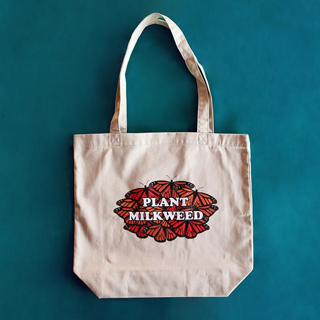 A khaki-colored tote bag with text saying "Plant Milkweed" on it. The text is surrounded by illustrations of red and orange monarch butterflies.