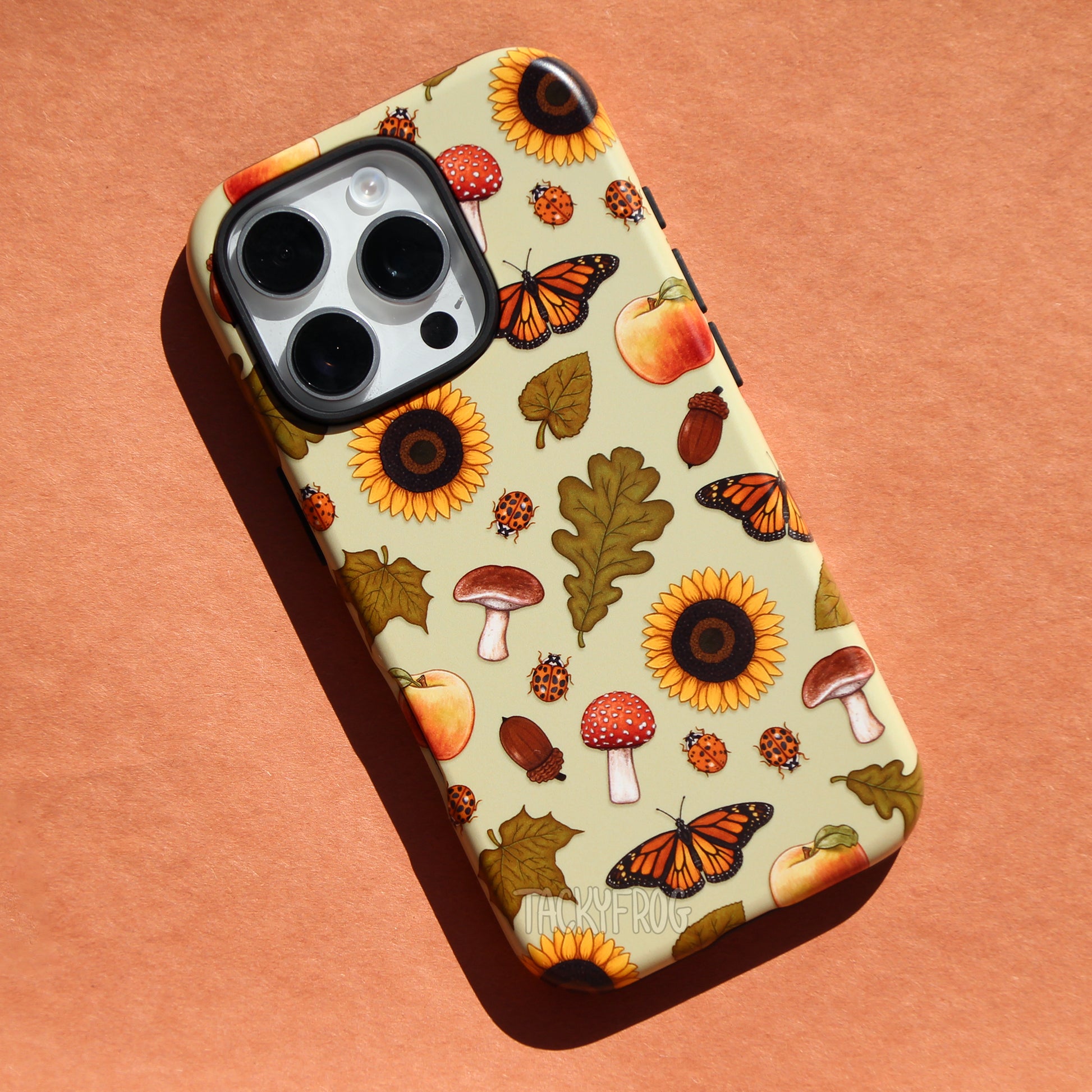Phone case with early fall-inspired design depicting sunflowers, leaves, mushrooms, ladybugs, apples, and butterflies against a very light green background.