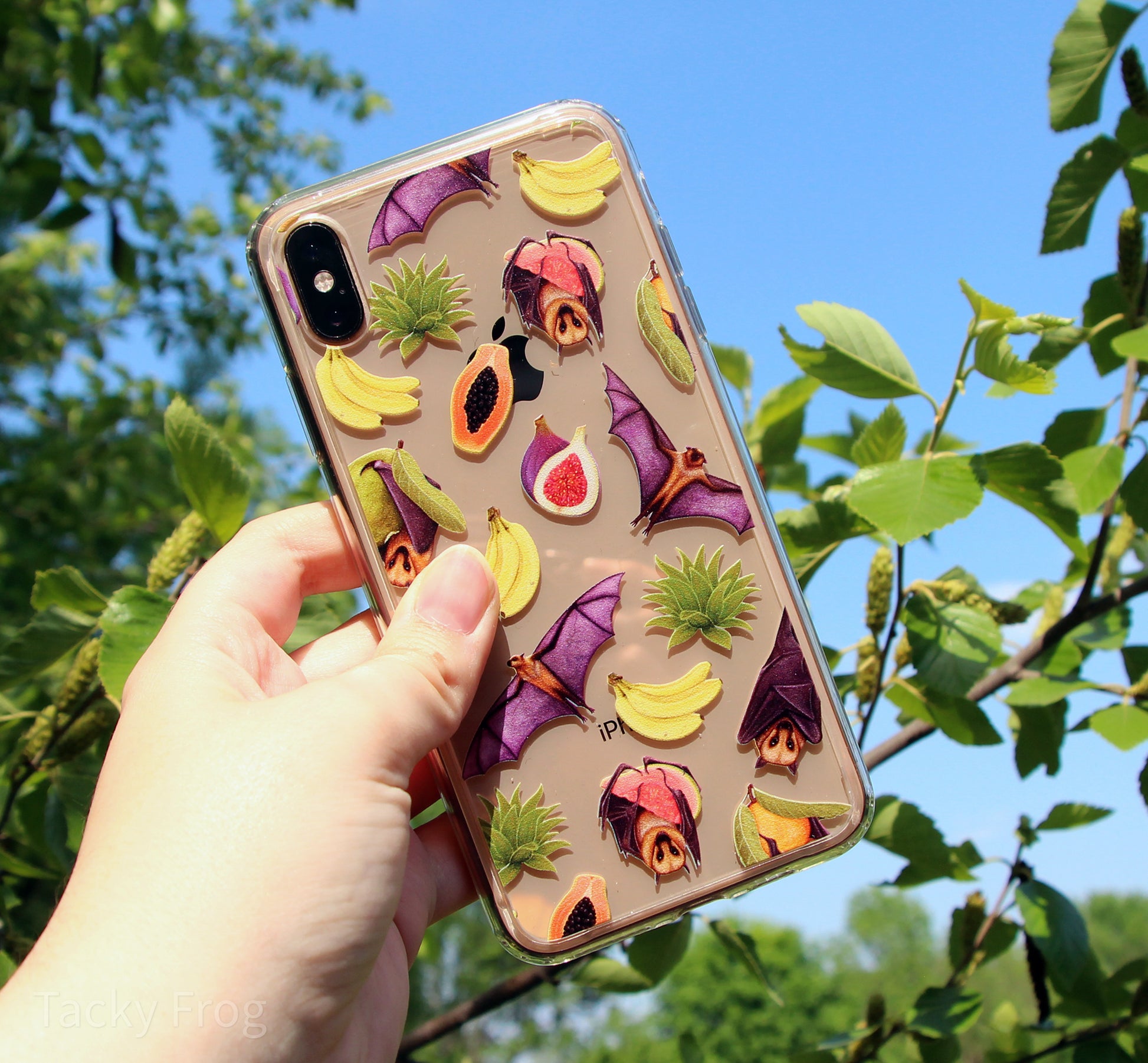 The clear fruit bat phone case held up in front of a tree.