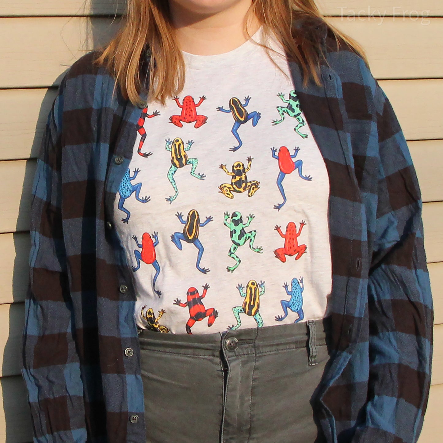 A woman wearing a colorful dart frog shirt under a blue flannel. The color variant she is wearing is "ash".