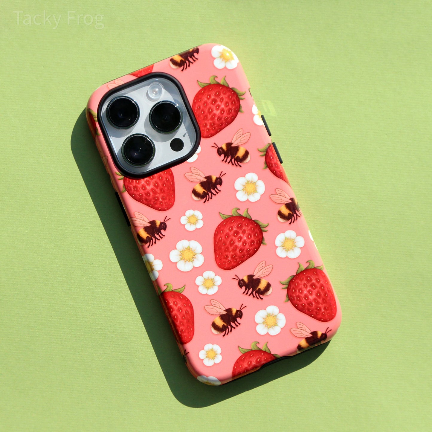 A pink phone case that has a pattern of strawberries, bumblebees, and white flowers on it.