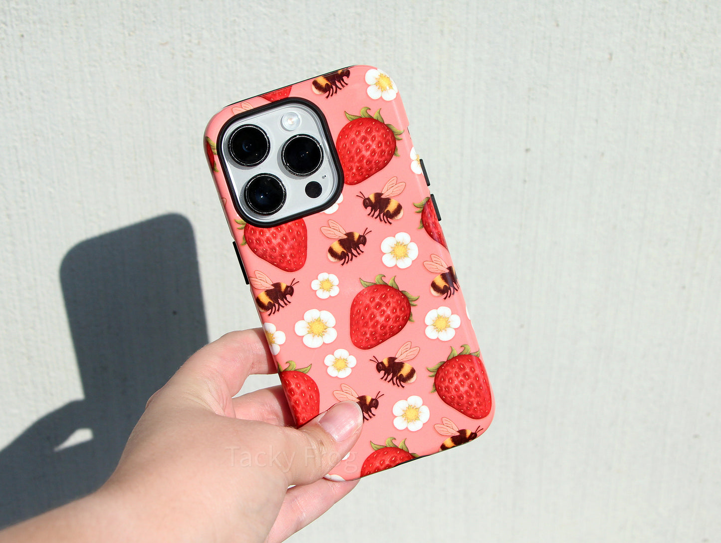 The strawberries and bees phone case held in front of a concrete wall. The case is dual-layered, with the inner layer being solid black.