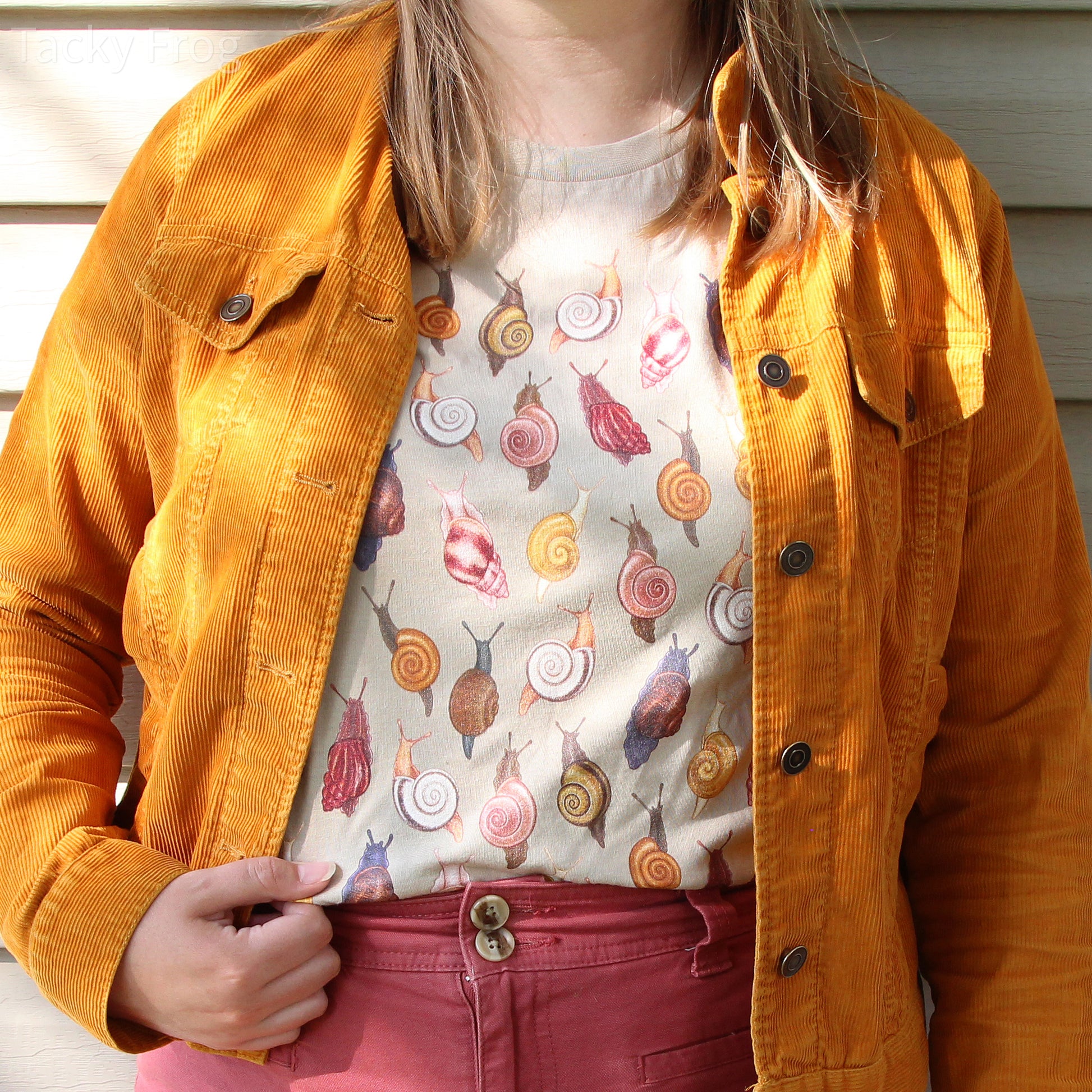 A woman wearing a snail t-shirt in the color "Tan" under a yellow jacket. The t-shirt is worn tucked-in. The shirt features digitally-drawn illustrations of various snails.