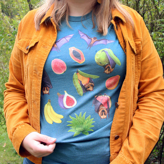 A woman wearing the fruit bat t-shirt in the color "Heather Deep Teal" under a yellow jacket. The design on the shirt features fruit bats with some of their foods, including guavas, bananas, agave, figs, and papaya.