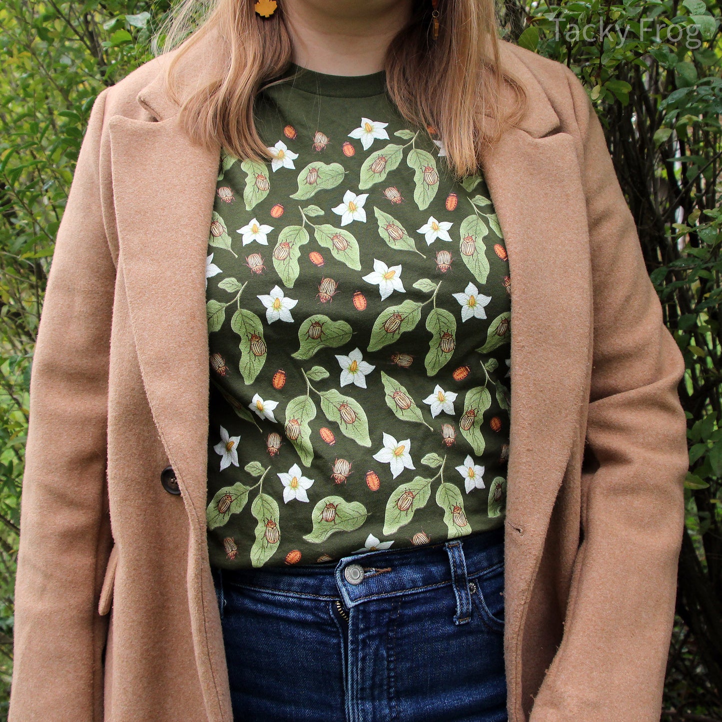 A woman wearing an olive green-colored t-shirt featuring a design of potato beetles and their larvae, potato leaves, and potato flowers. She is wearing the shirt under a long tan coat.