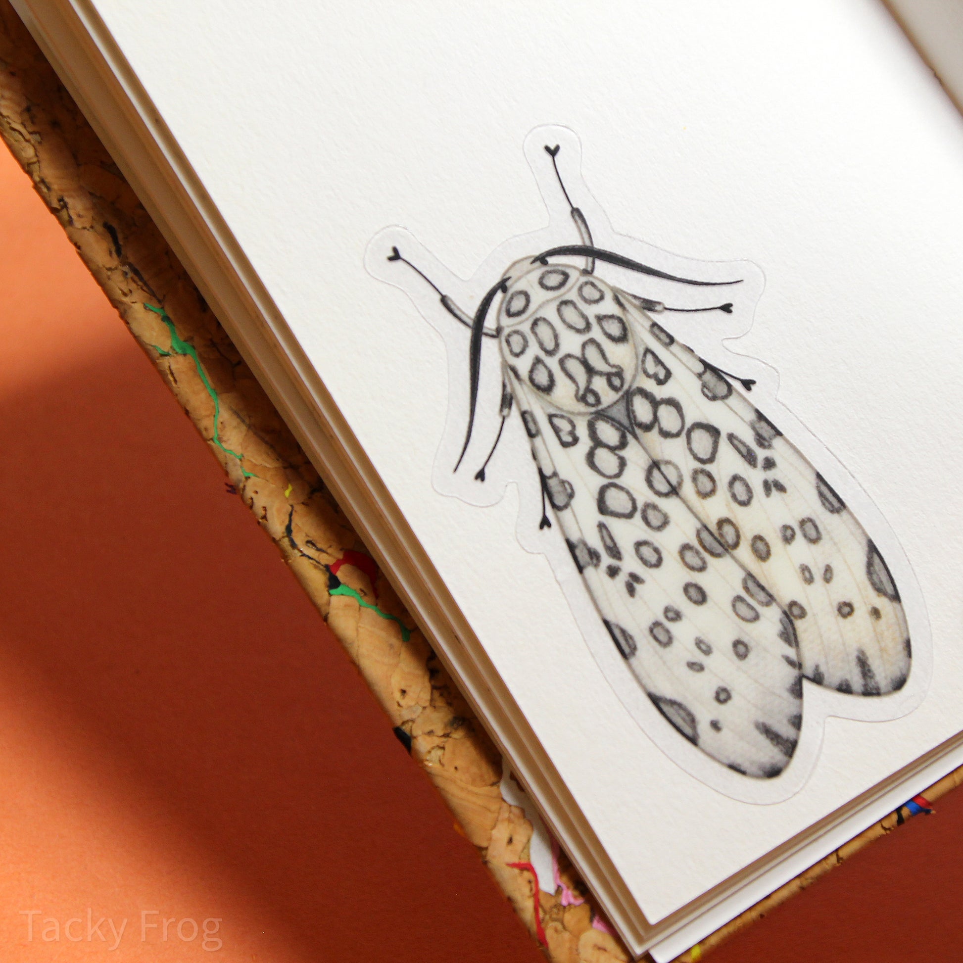 The leopard moth clear vinyl sticker placed on a piece of blank paper inside of a sketchbook.