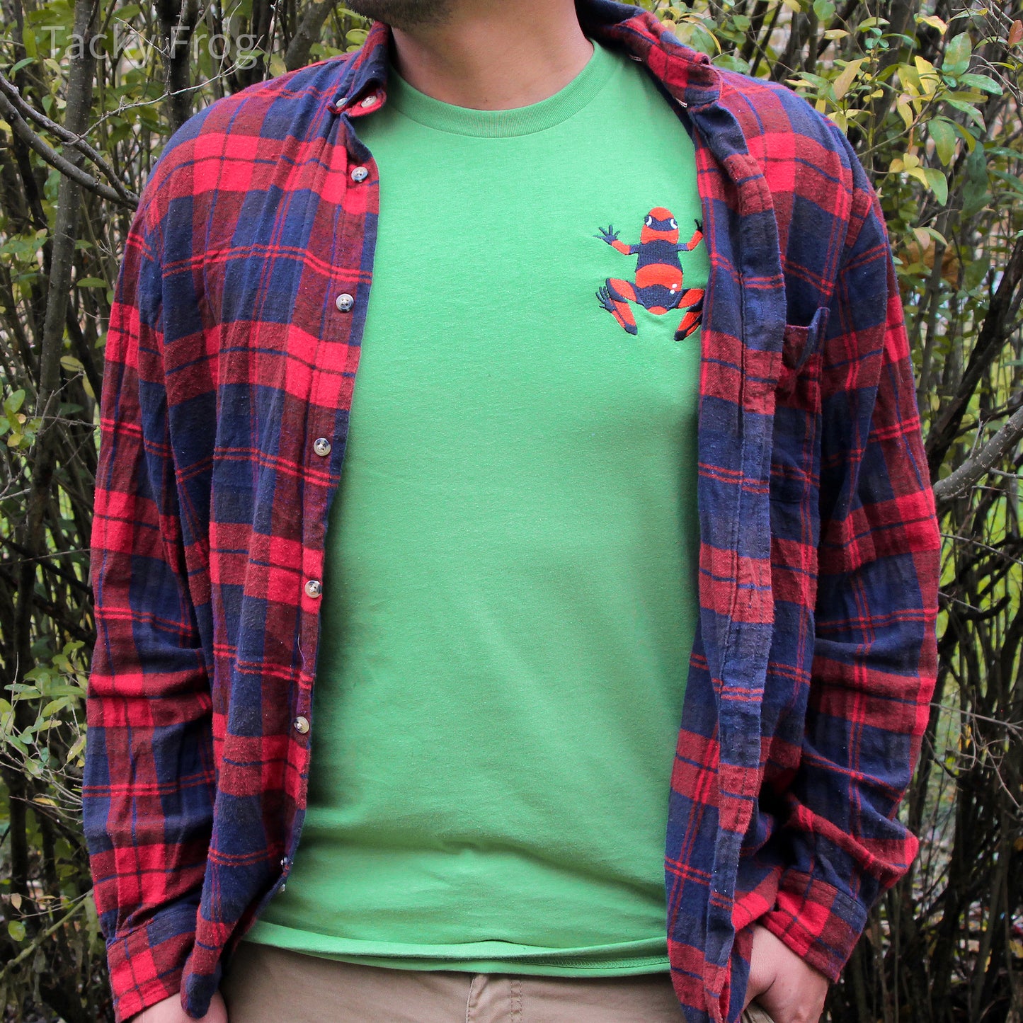 A man wearing the red dart frog embroidered t-shirt in the color "leaf" under a red and blue flannel.