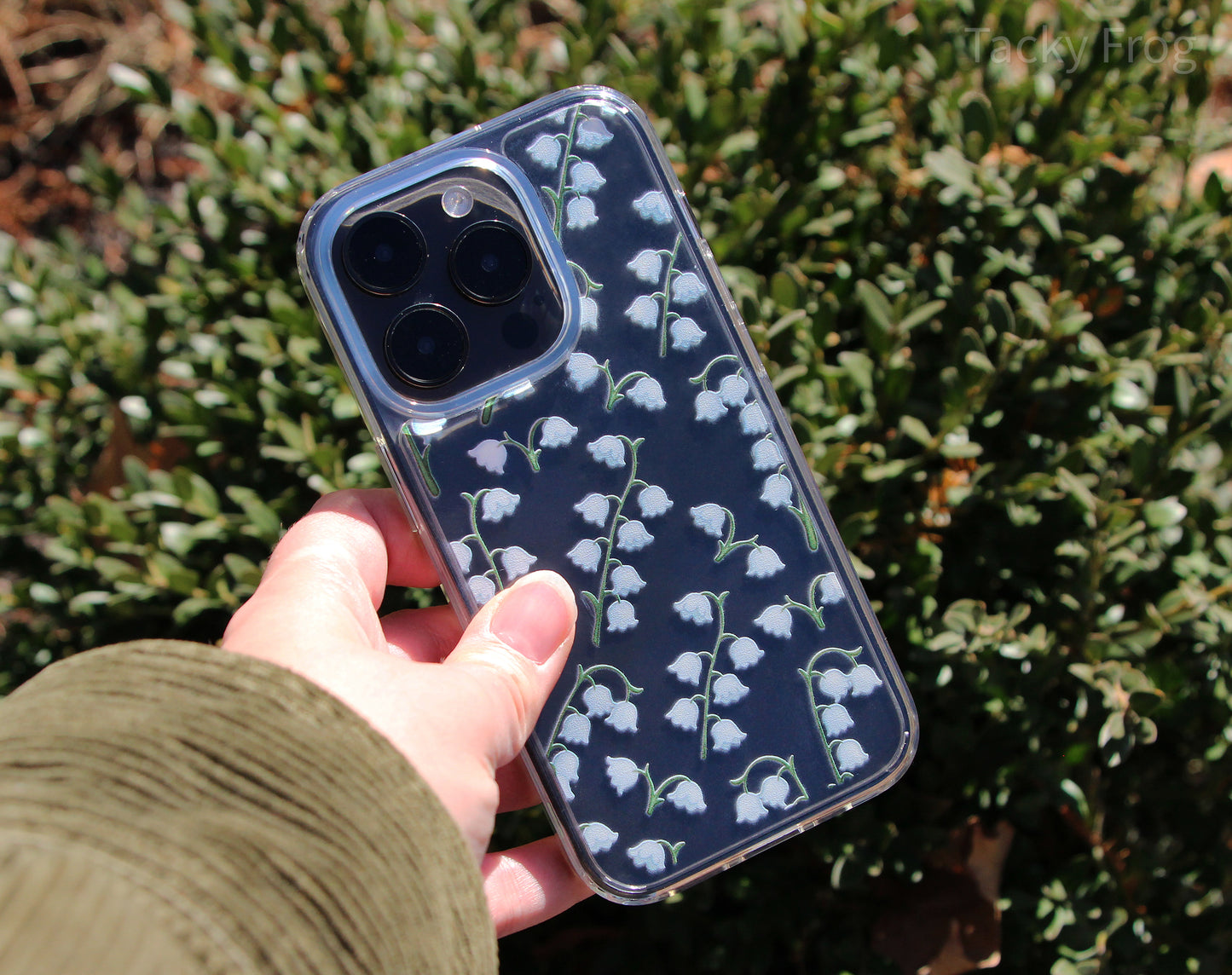 The lily of the valley-patterned phone case held in front of a bush.