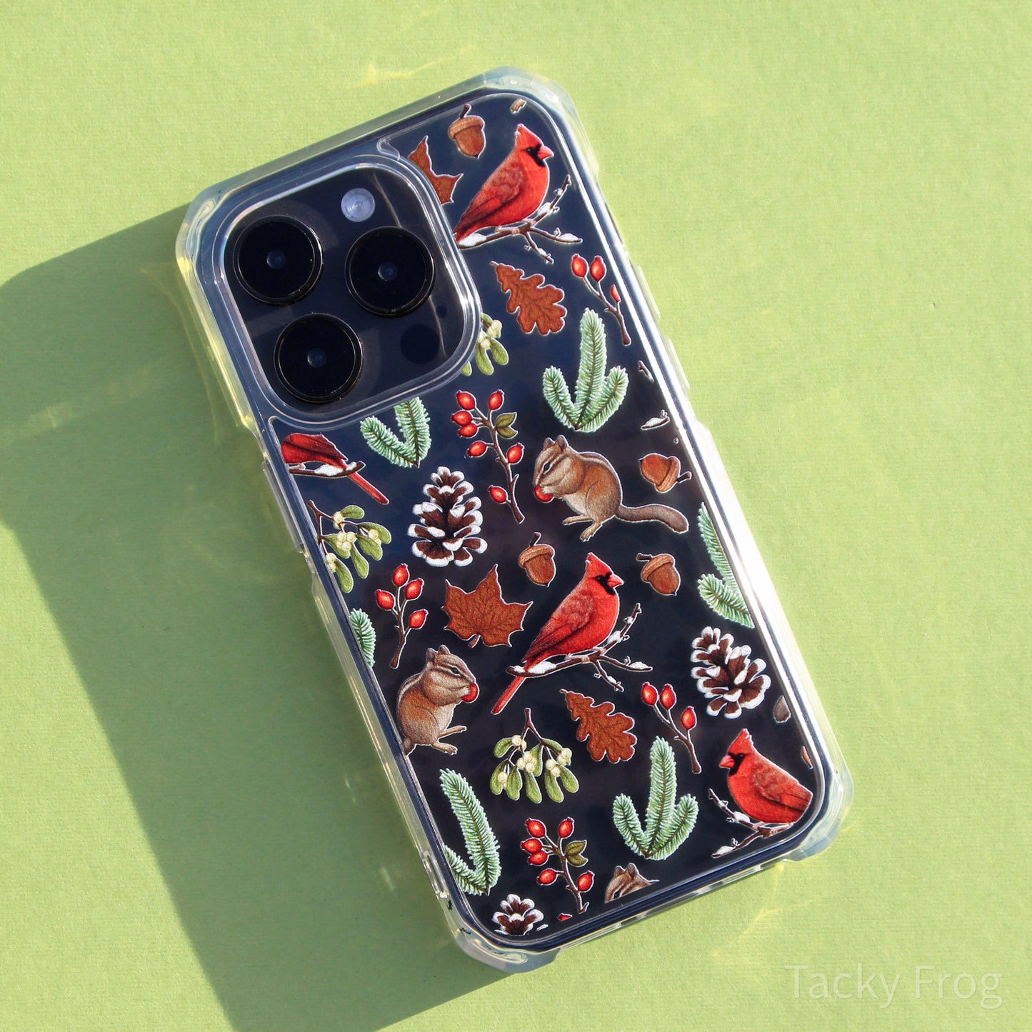 A clear vinyl sticker featuring a pattern of winter nature walk-inspired elements, including red cardinals, chipmunks, snowy pinecones, rose hip, mistletoe, pine leaves, and more. it is placed on a dark-colored phone.