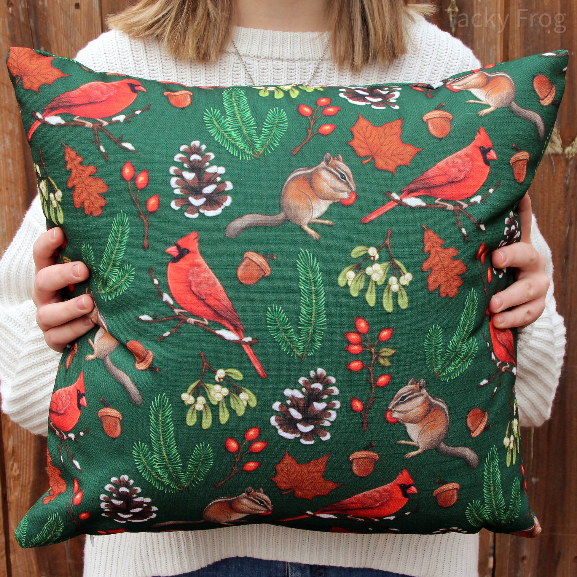 An 18 x 18 inch dark green throw pillow featuring a pattern of various winter-inspired animals and plants, including chipmunks, red cardinals, snowy pinecones, mistletoe, and more.