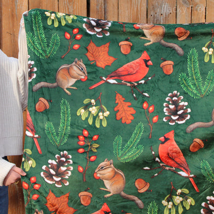 A winter nature hike-inspired throw blanket held up in the air. The pattern features illustrations of cardinals, chipmunks, snowy pinecones, pine needles, and more.