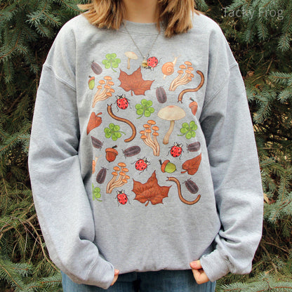 A grey sweatshirt featuring a design made up of various nature-inspired elements, including snails, millipedes, pill bugs, mushrooms, ladybugs, clovers, acorns, and dead leaves.