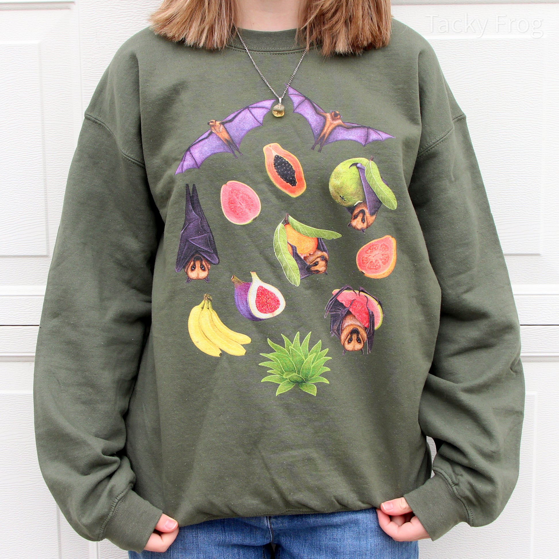 A young woman wearing a military green sweatshirt featuring a design of fruit bats and tropical fruits.