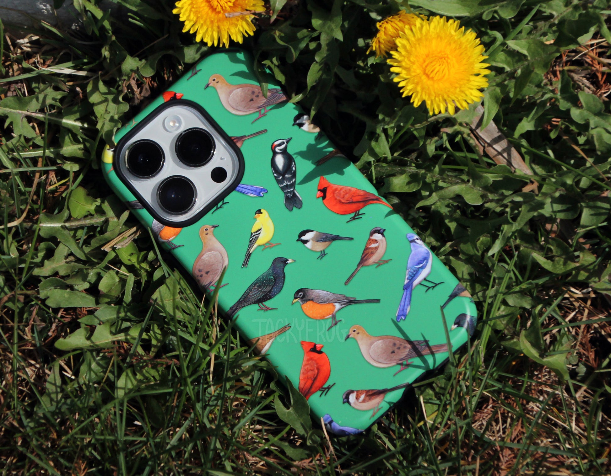 The bird-patterned tough phone case placed on the grass, next to dandelions.