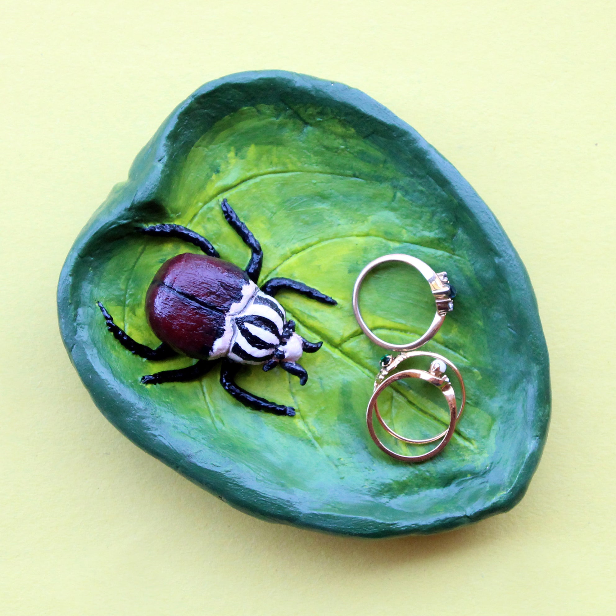 A clay trinket tray that looks like a Goliath beetle on a leaf. The tray was hand-painted and hand-sculpted. There are three rings in the tray for scale.