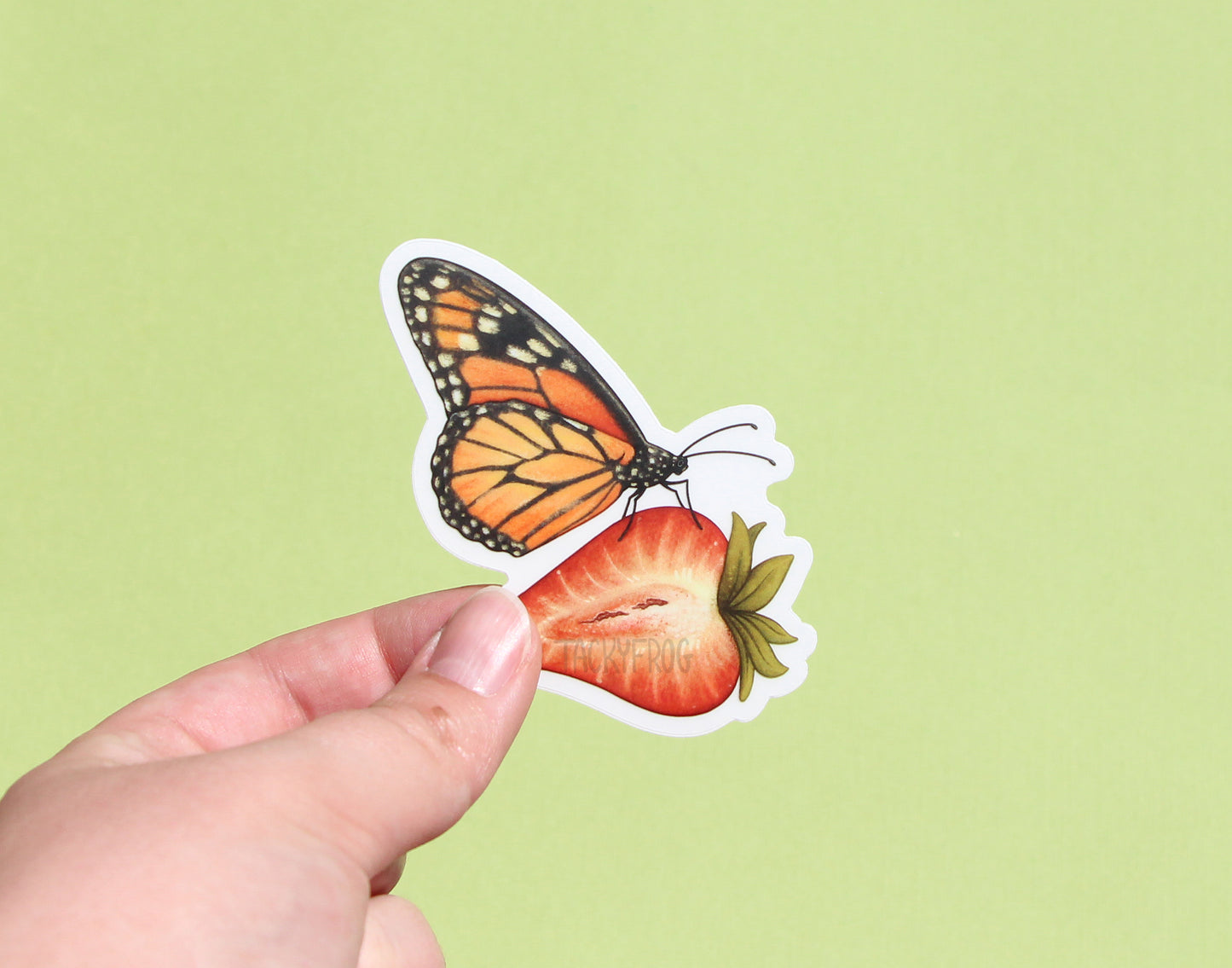 A clear vinyl sticker of a monarch butterfly on a strawberry with the white paper backing still on.