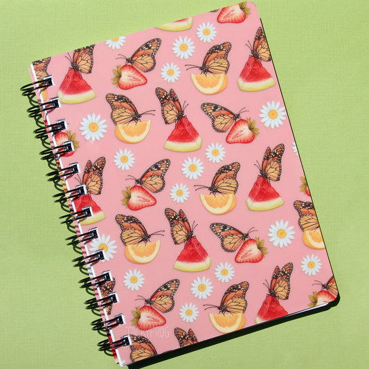 A light pink notebook featuring a pattern of daisies and monarch butterflies with strawberries, lemons, and watermelon.