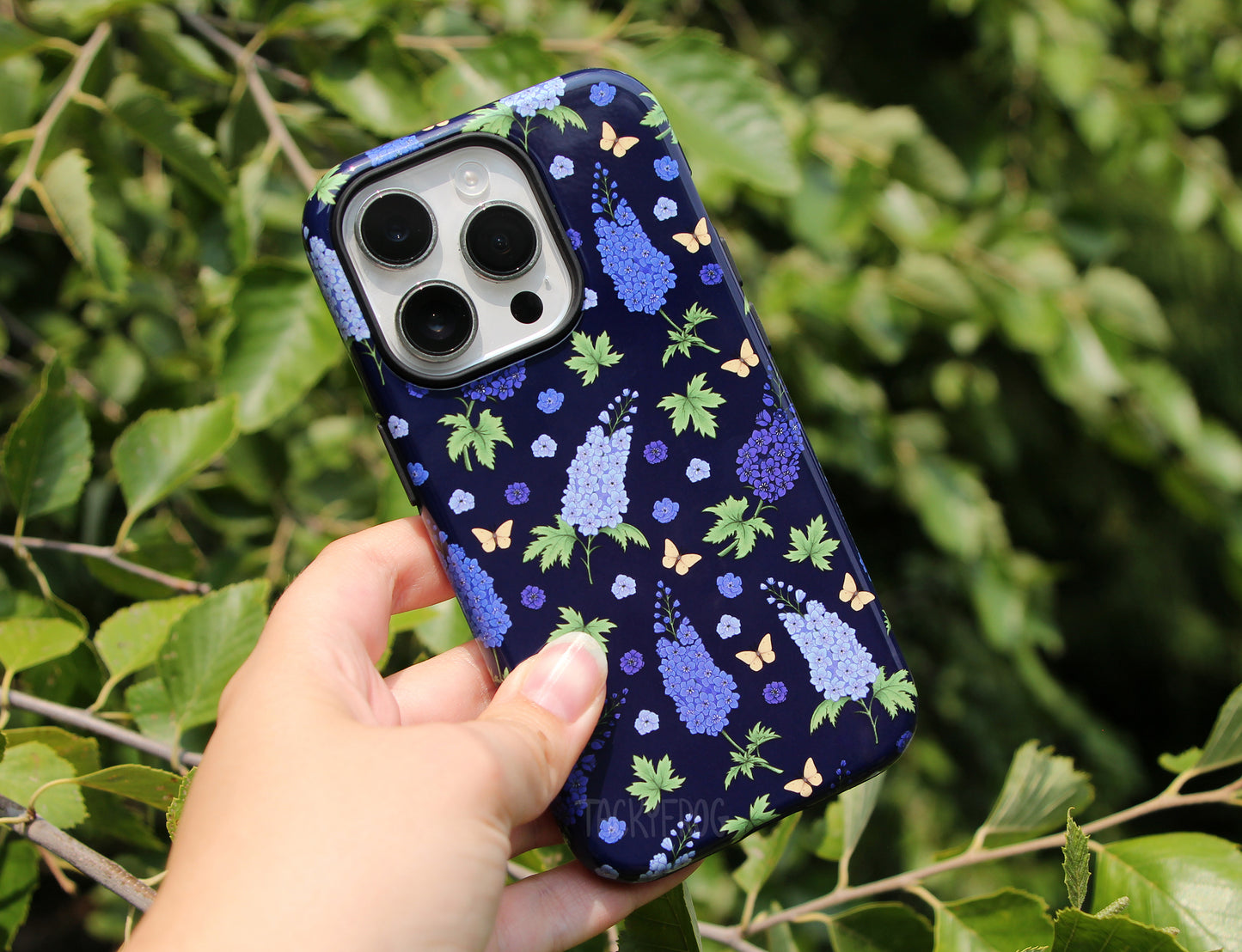 The blue Larkspur phone case held up in front of a tree.