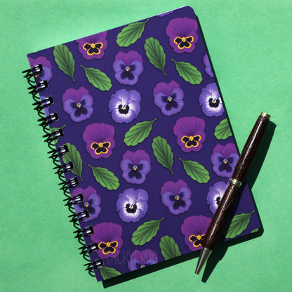 A 5 x 7 inch purple notebook featuring a pattern of purple pansies and leaves. The pen is not included.