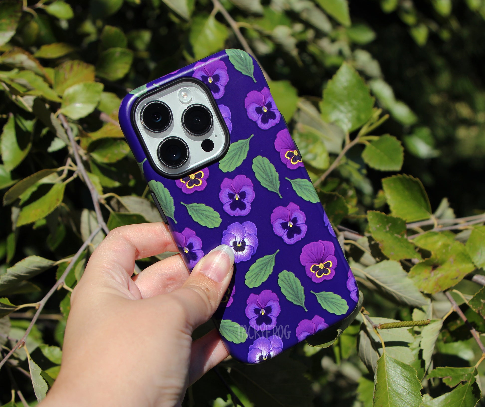 The purple pansies phone case shown in front of a tree.