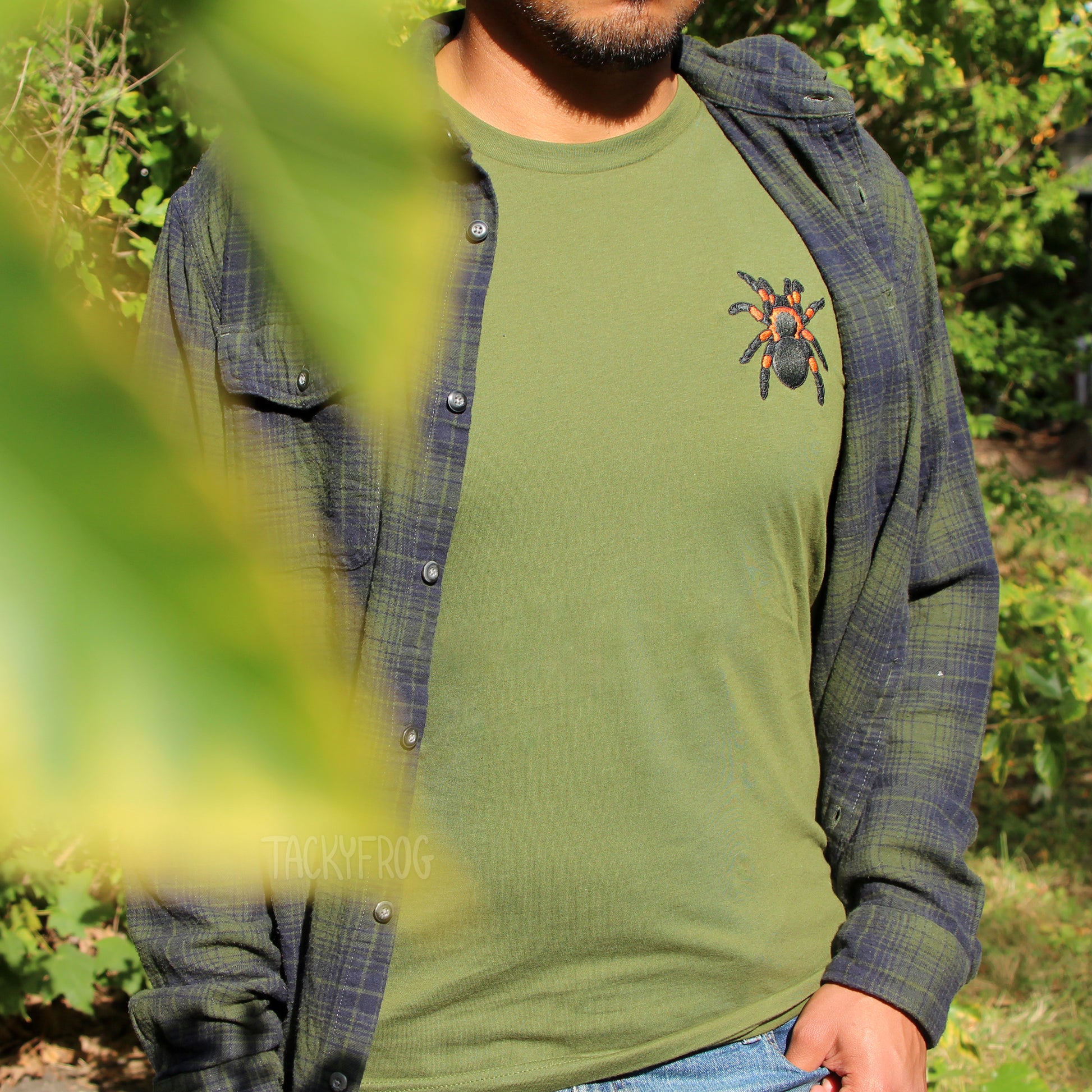 A man wearing the red-knee tarantula embroidered shirt in the olive green color under a dark green flannel. A couple of leaves partially hide him from view.