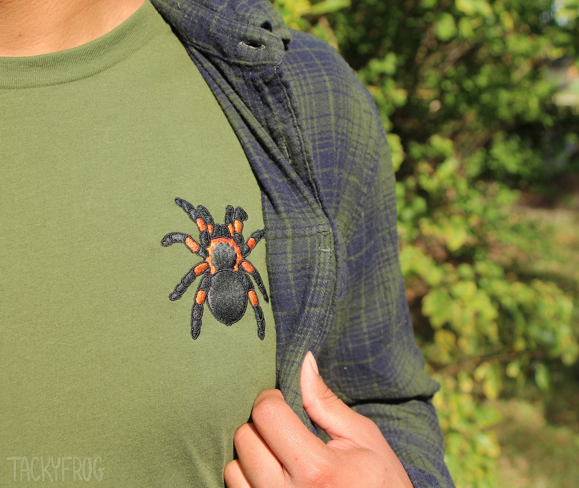 A closeup of the machine-embroidered details on the tarantula-embroidered shirt. A dark green flannel is worn over the shirt. 