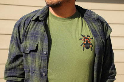 A man wearing the embroidered red-knee tarantula t-shirt in the color "olive" under a green flannel.