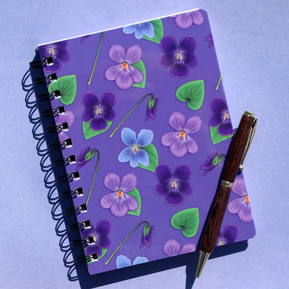 A purple 5x7 inch mini spiral-bound, ruled-line notebook featuring a pattern of green leaves and violet flowers. The pen is not included.