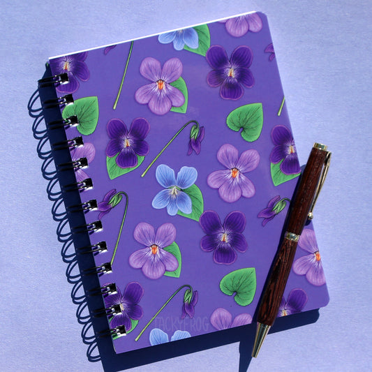 A purple 5x7 inch mini spiral-bound, ruled-line notebook featuring a pattern of green leaves and violet flowers. The pen is not included.