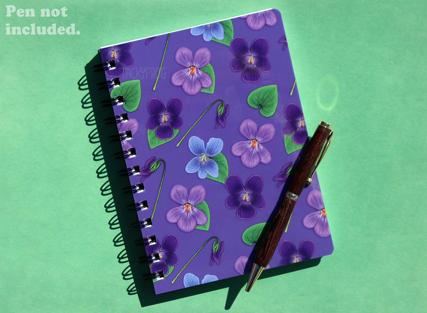 Another view of the violet notebook, this time against a green background. The text in the upper left corner reads "pen not included".