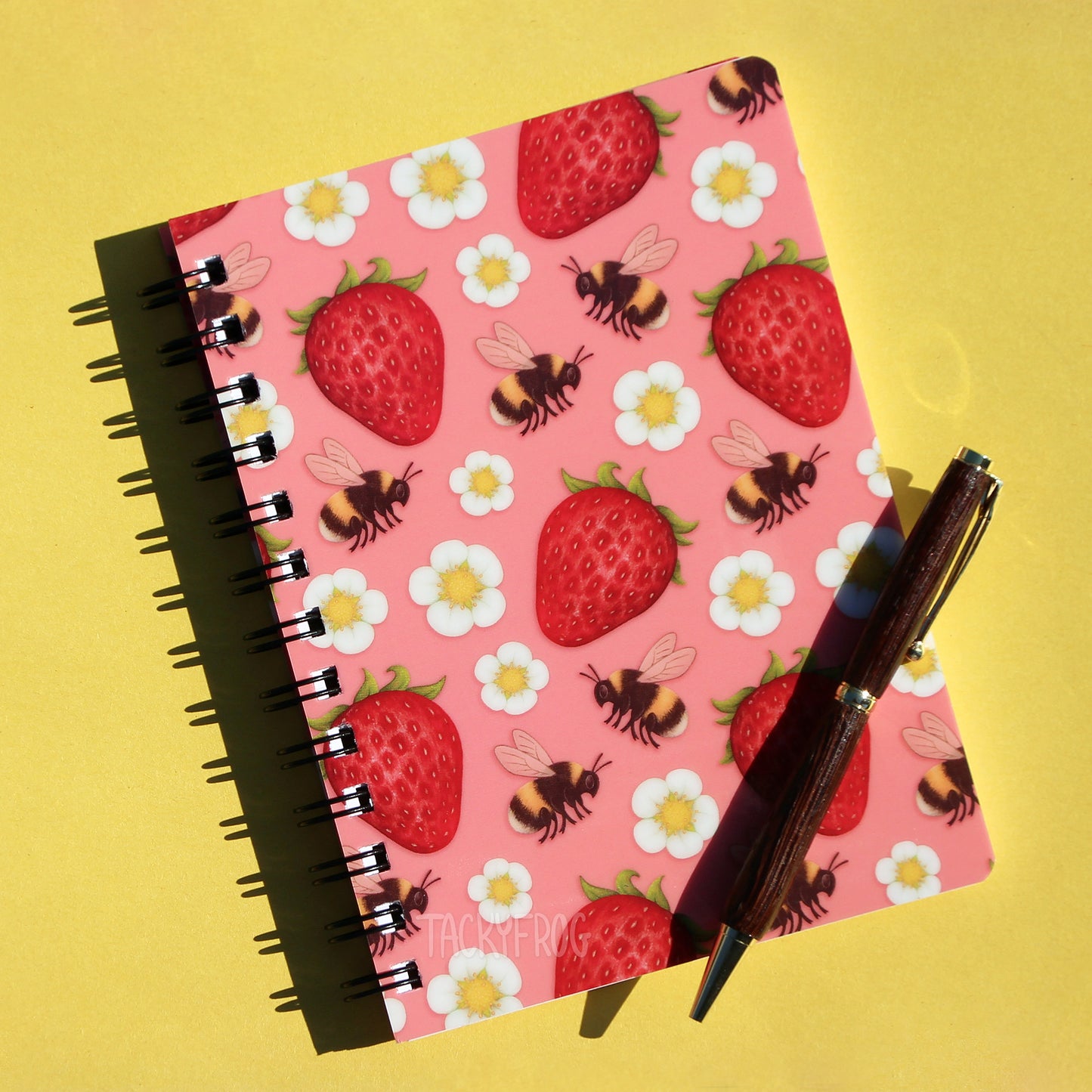 A pink 5x7 inch mini spiral-bound, ruled-line notebook featuring a pattern of bumblebees, strawberries, and white and yellow flowers. The pen is not included.