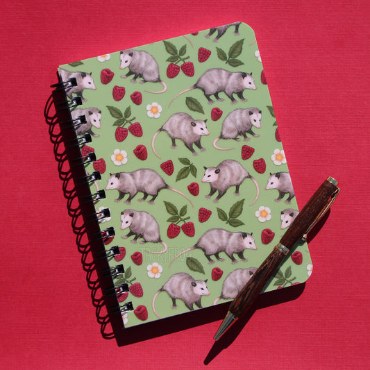 A 5 by 7 inch mini green notebook featuring a pattern of opossums, raspberries, leaves, and white flowers. The pen is not included.