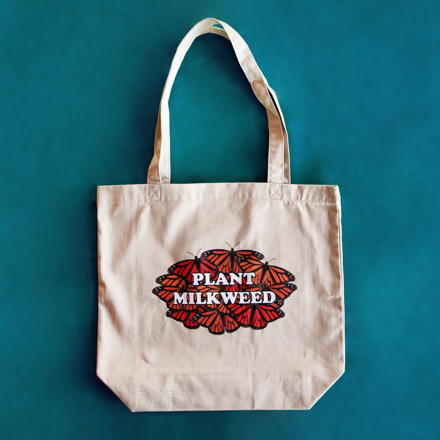 A tan-colored tote bag featuring a design of several monarch butterflies underneath text reading "Plant Milkweed". 