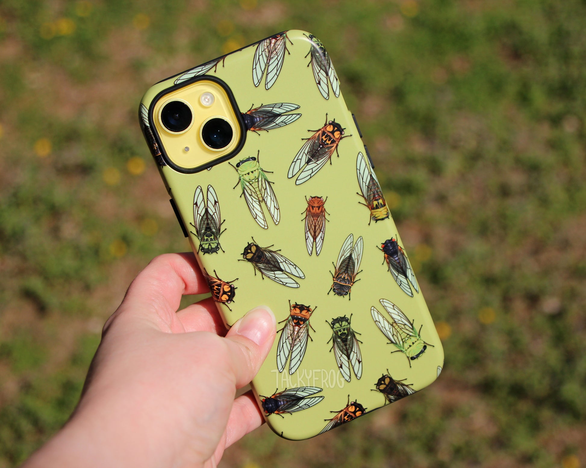 A cicada-patterned phone case, shown on a yellow phone, held above grass.