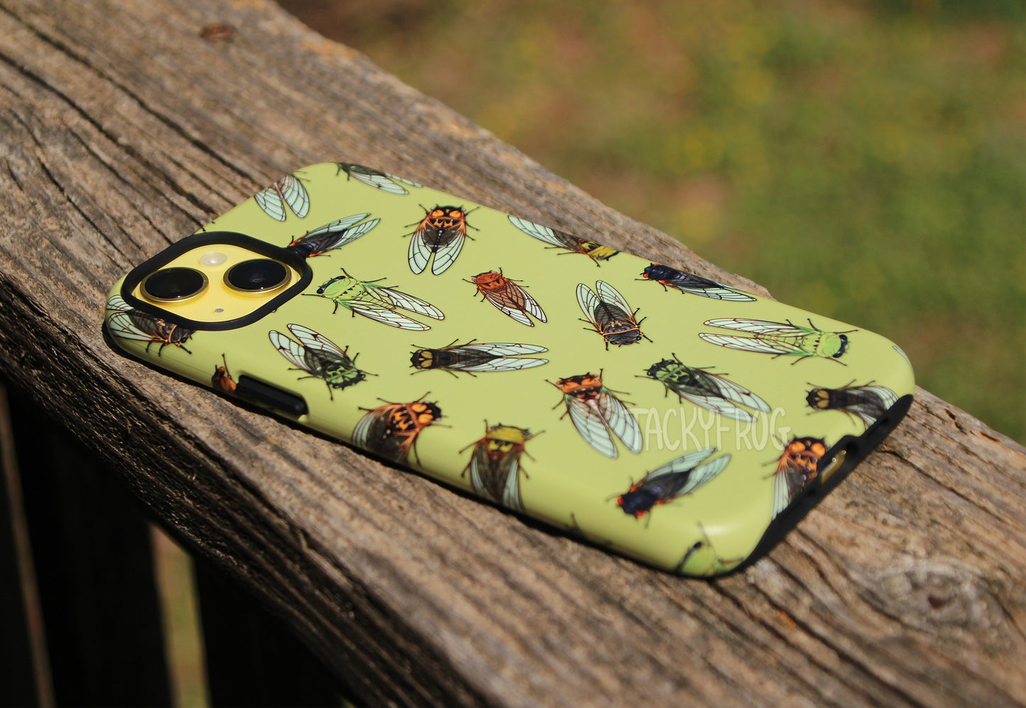 An angled view of the cicada-patterned phone case, shown on a yellow phone and placed on wooden deck railing. 