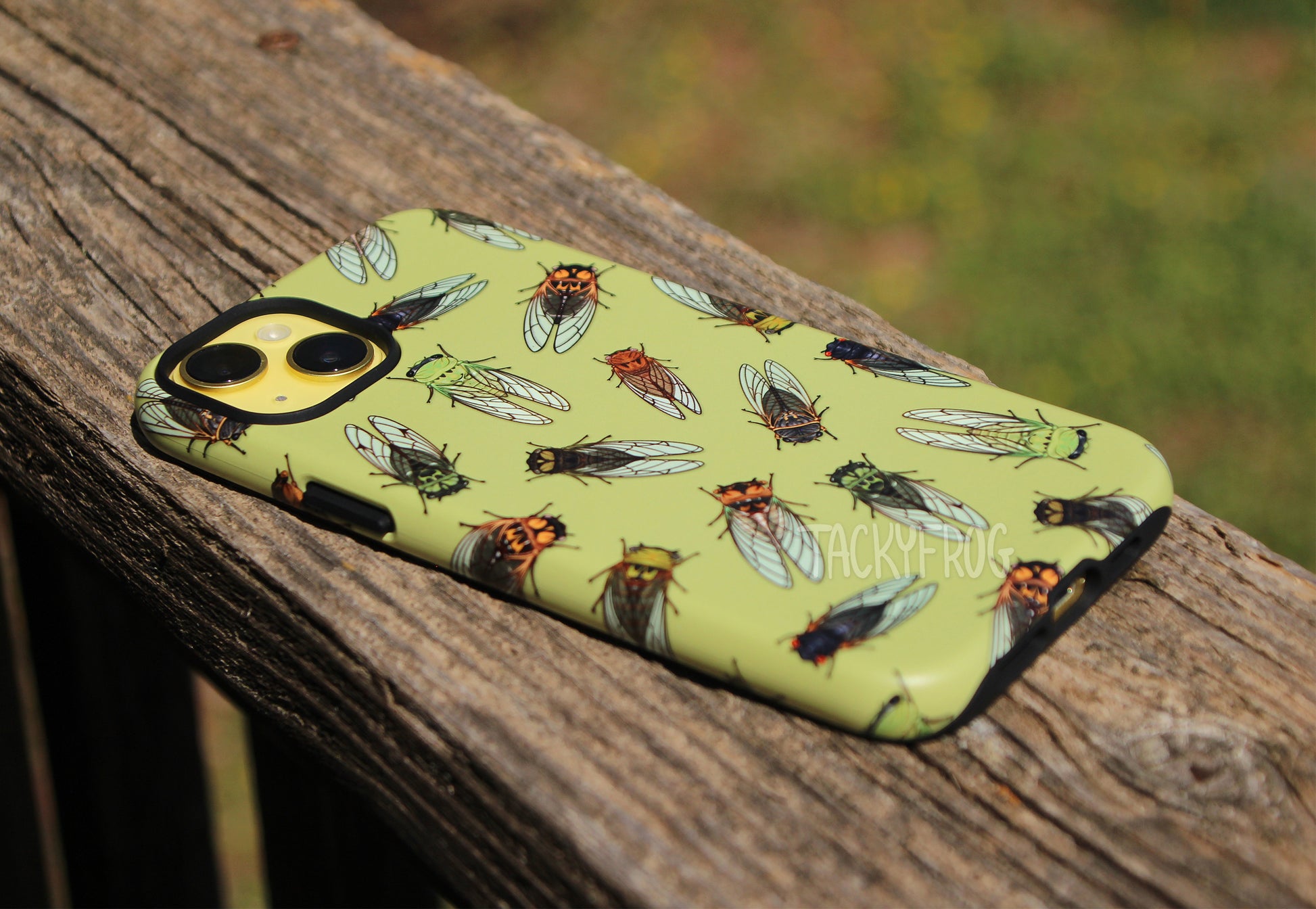An angled view of the cicada-patterned phone case, shown on a yellow phone and placed on wooden deck railing. 