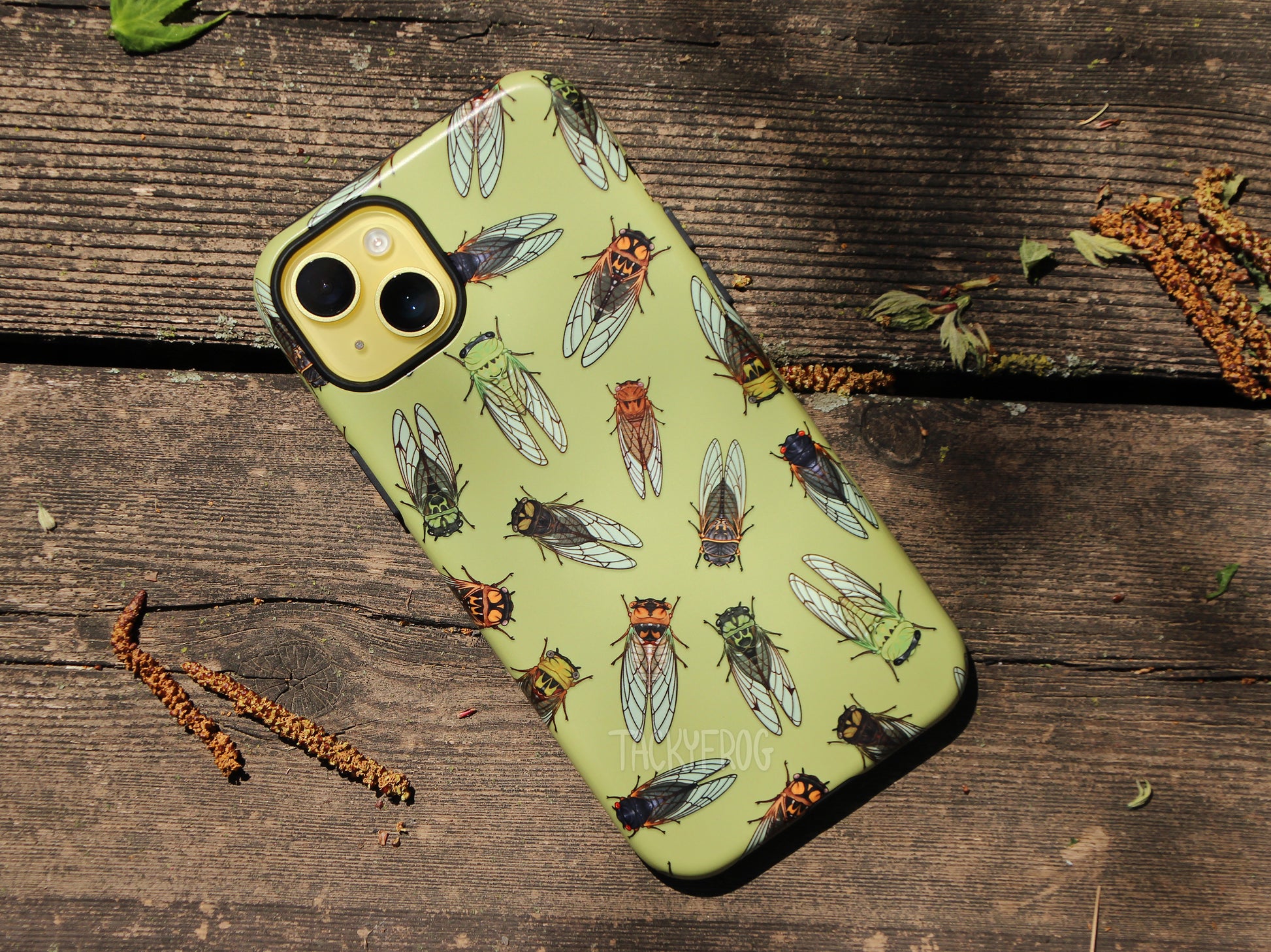 A light green phone case featuring a pattern of cicadas, shown on a yellow phone, placed on a wooden deck.
