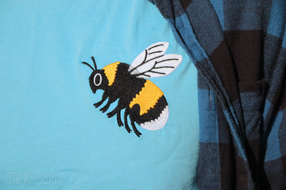 Close up of the embroidered bee design on the ocean blue t-shirt. It is worn under a black and blue flannel which is not included.