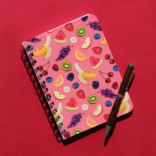 A pink 5 x 7 inch spiral bound mini journal featuring a pattern of apple slices, strawberries, blueberries, raspberries, blackberries, orange slices, watermelon slices, kiwi, bananas, cherries, and grapes. The pen is not included.