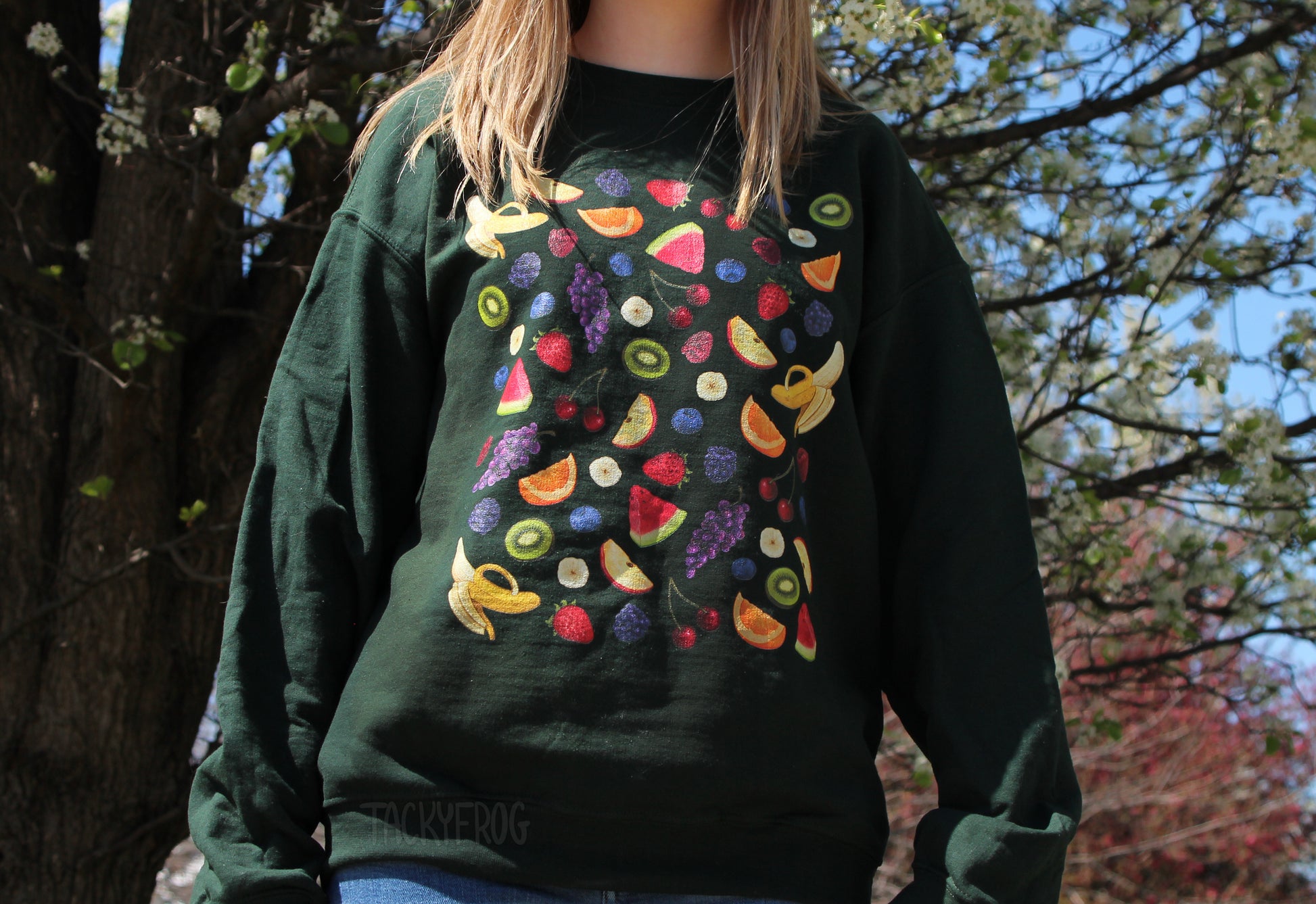 A woman standing near a tree while wearing the fruit salad sweatshirt in the color "forest green"