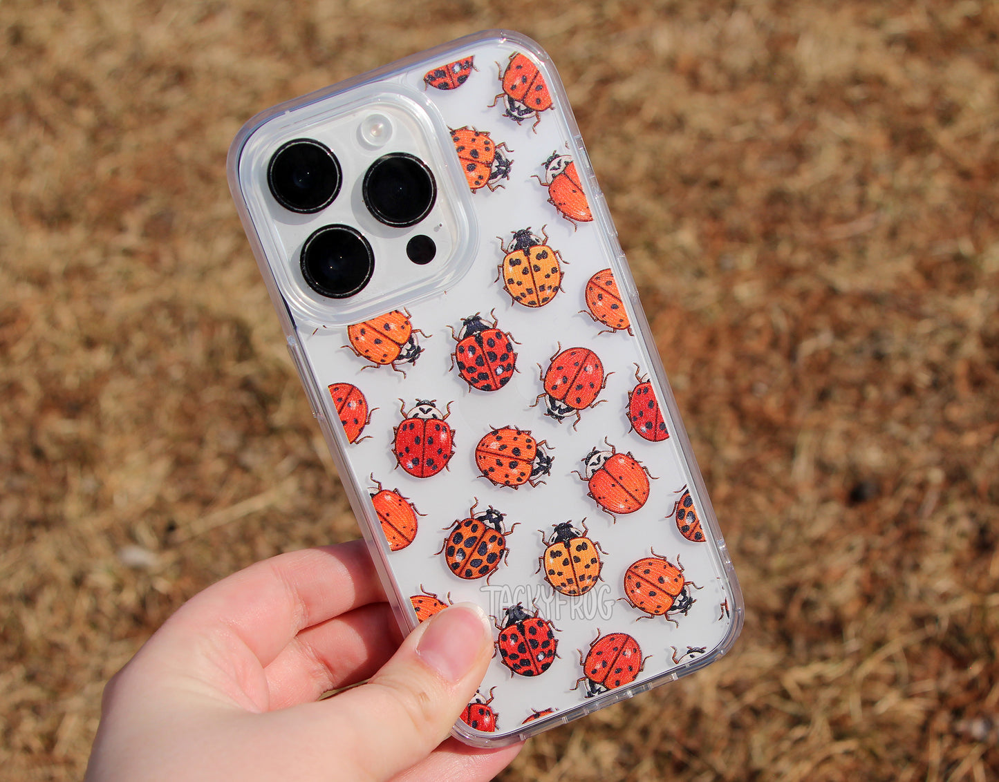 The ladybugs clear phone case held over dead grass. It is shown on a light-colored phone.