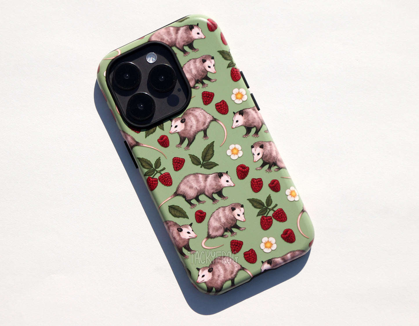 A green phone case featuring a pattern of opossums, raspberries, green leaves, and white flowers. The case is shown on a dark grey phone.
