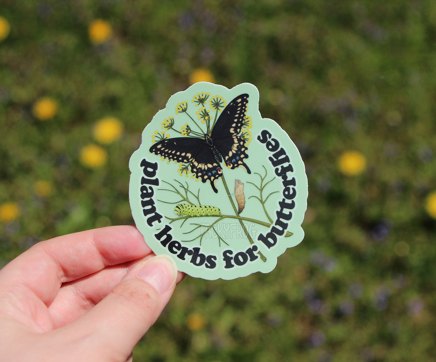 Sticker with butterfly design and text that reads "Plant herbs for butterflies" held above summer vegetation.