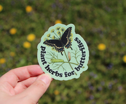 Sticker with butterfly design and text that reads "Plant herbs for butterflies" held above summer vegetation.