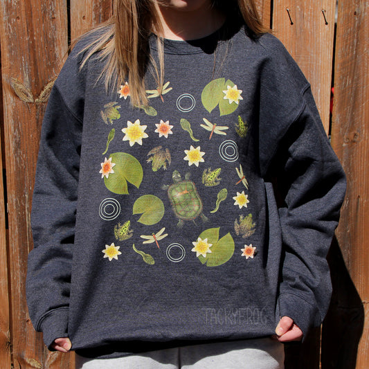A woman wearing a dark grey sweatshirt featuring a design inspired by pond life. A turtle, frogs, dragonflies, tadpoles, water lilies, and lily pads can be seen on the sweatshirt. The sweatshirt color is "dark heather".