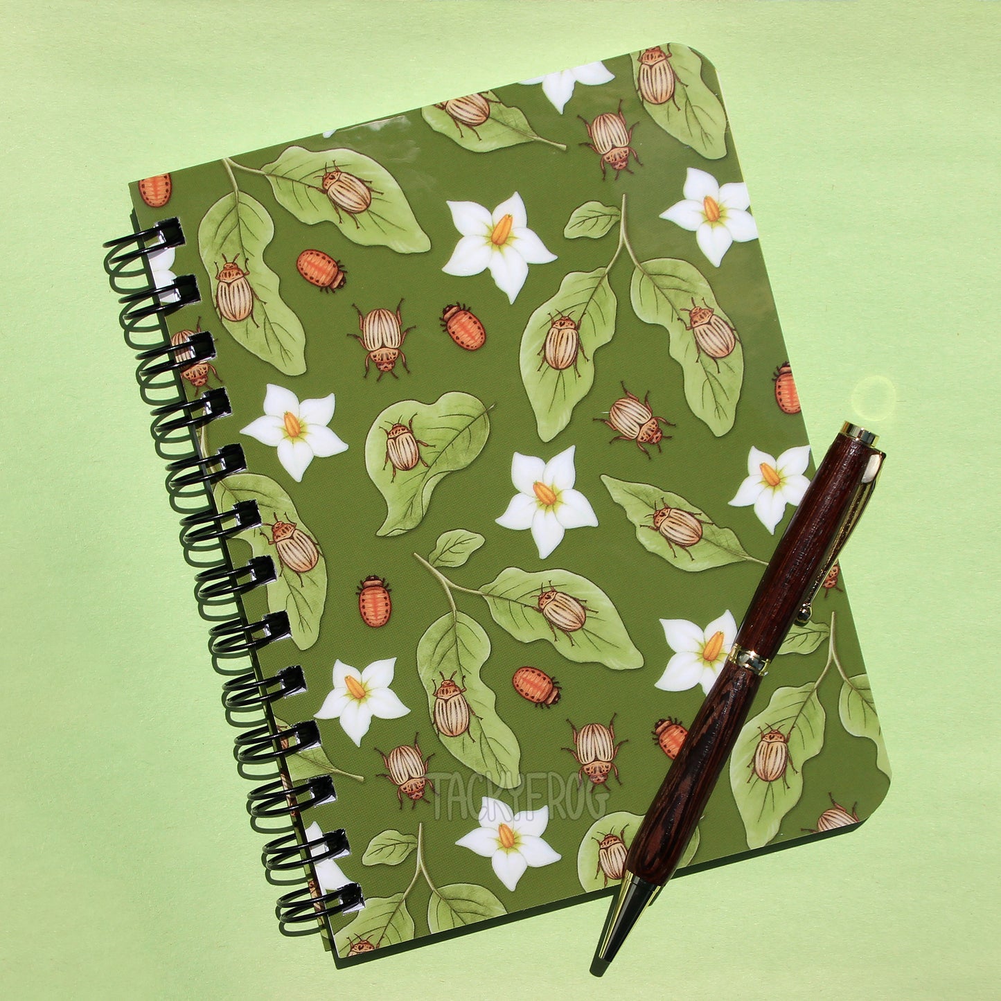 Notebook featuring a pattern of potato beetles, light green leaves, white flowers, and potato beetle larvae. The pen is not included.